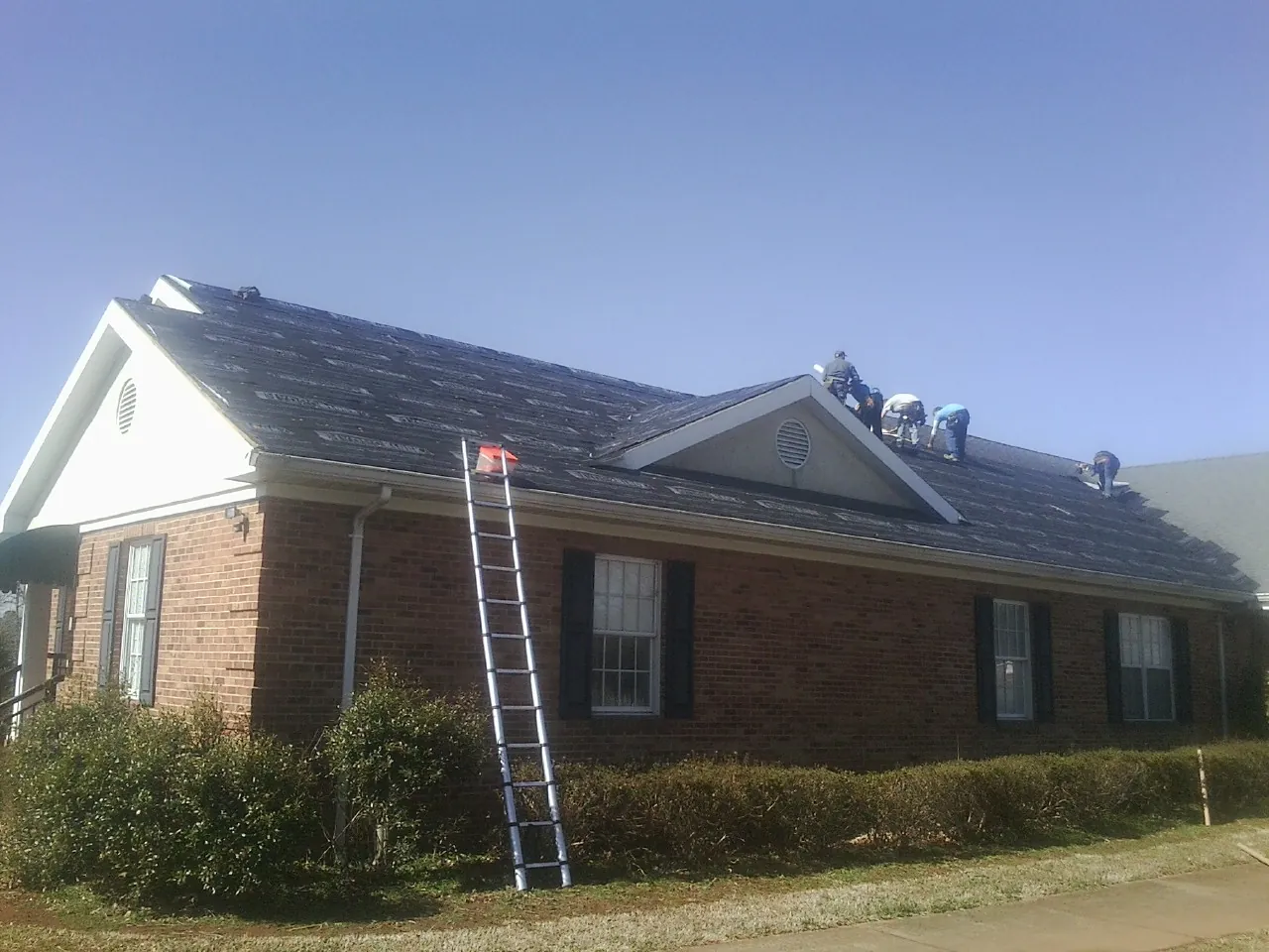 Crew tearing shingles off a brick ranch roof with a ladder set — Emergency Roof Repair work by our crew in Dameron