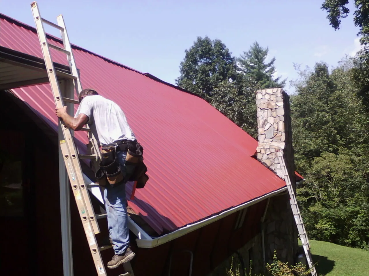 Roofer on a ladder beside a red metal roof and stone chimney — Soffit & Fascia Repair by Roofing Brothers in Dameron